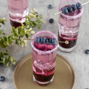3 blueberry gin cocktails with a sugared rim and topped with a cocktail pin of blueberries.