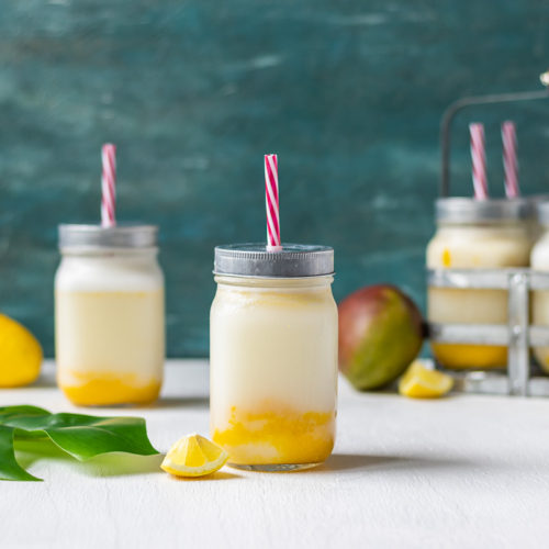 Creamy Mango Lemonade in a mason jar with a red and white straw. In the background is more lemonades, stacks of lemons, a mango, and a tropical leaf.