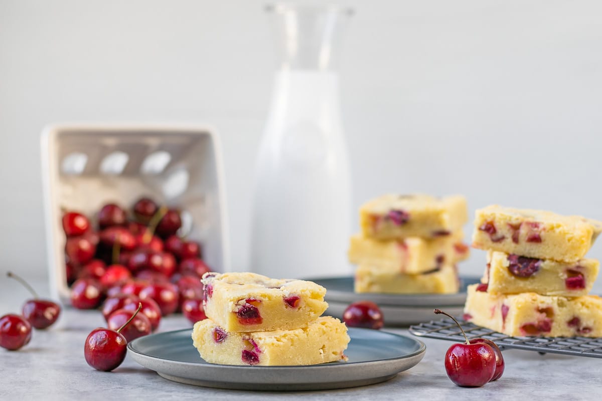 Cherry White Chocolate Blondies- stack of two surrounded by other stacks of blondies, fresh cherries, and a container of milk.