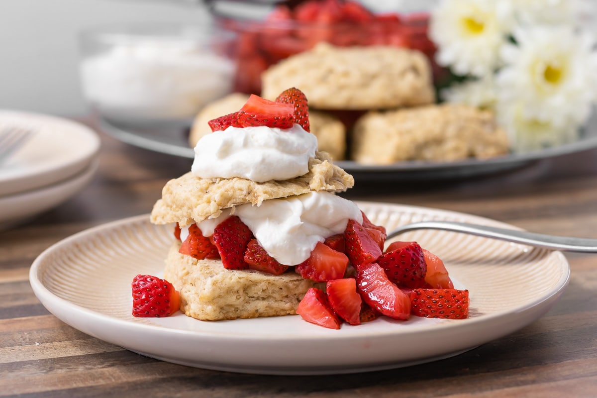 Strawberry Banana Shortcakes assembled and sitting on a decorative plate. Layers of shortcake, fresh strawberry syrup, and whipped cream. Ingredients in background.