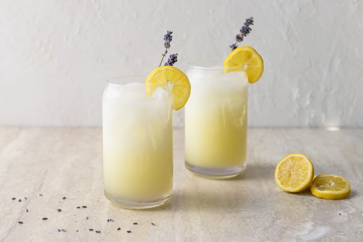 A pair of Lavender Coconut Lemonades. Shows the beautiful layers of white Coconut Milk, purple Lavender Simple Syrup, and yellow Lemonade as well as decorations of lemons and fresh lavender.