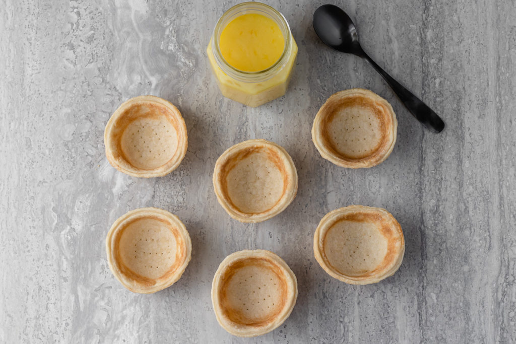 Overhead shot of empty mini pie crusts. Lemon curd set aside with a spoon.
