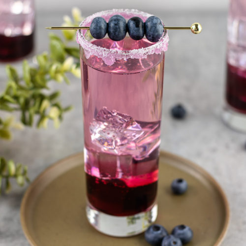 Layers of blueberry simple syrup, gin, and tonic water topped with blueberries on a cocktail stick. 3 tall glasses of the cocktail with greenery and fresh blueberries as decoration.