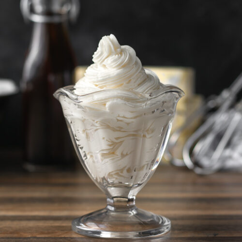 Frosting piped into a clear glass with bowl, vanilla, butter, and whisk in background.