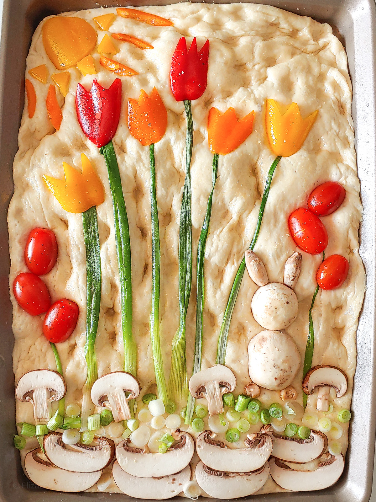Decorated Easter No Knead Focaccia bread. Bell peppers carved into tulips and sun, mushrooms made into bunnies and ground. chopped green onions as grass and stems.