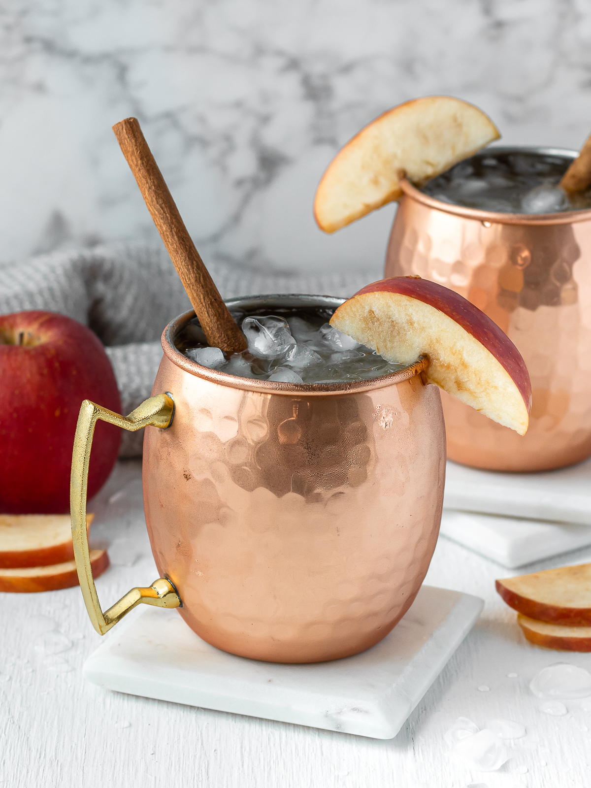 Two Apple Cider Mule Mocktails garnished with a cinnamon stick and slice of apple. More apples in the background.