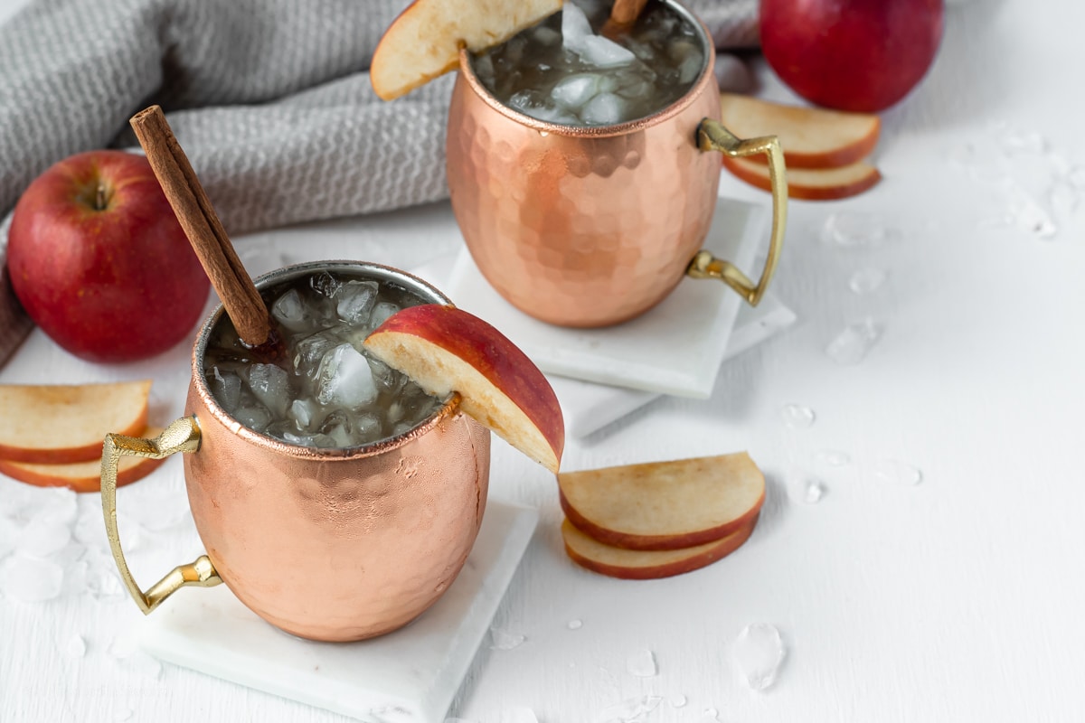 two apple cider mule mocktails with slices of apple cinnamon sticks in copper mugs surrounded by crushed ice