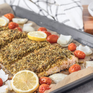 A fully baked pesto crusted salmon with roasted onions and tomatoes on the slide. Garnished with fresh lemon slices.