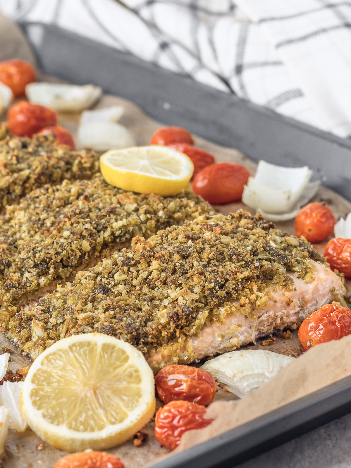 A fully baked pesto crusted salmon with roasted onions and tomatoes on the slide. Garnish of fresh lemon slices.