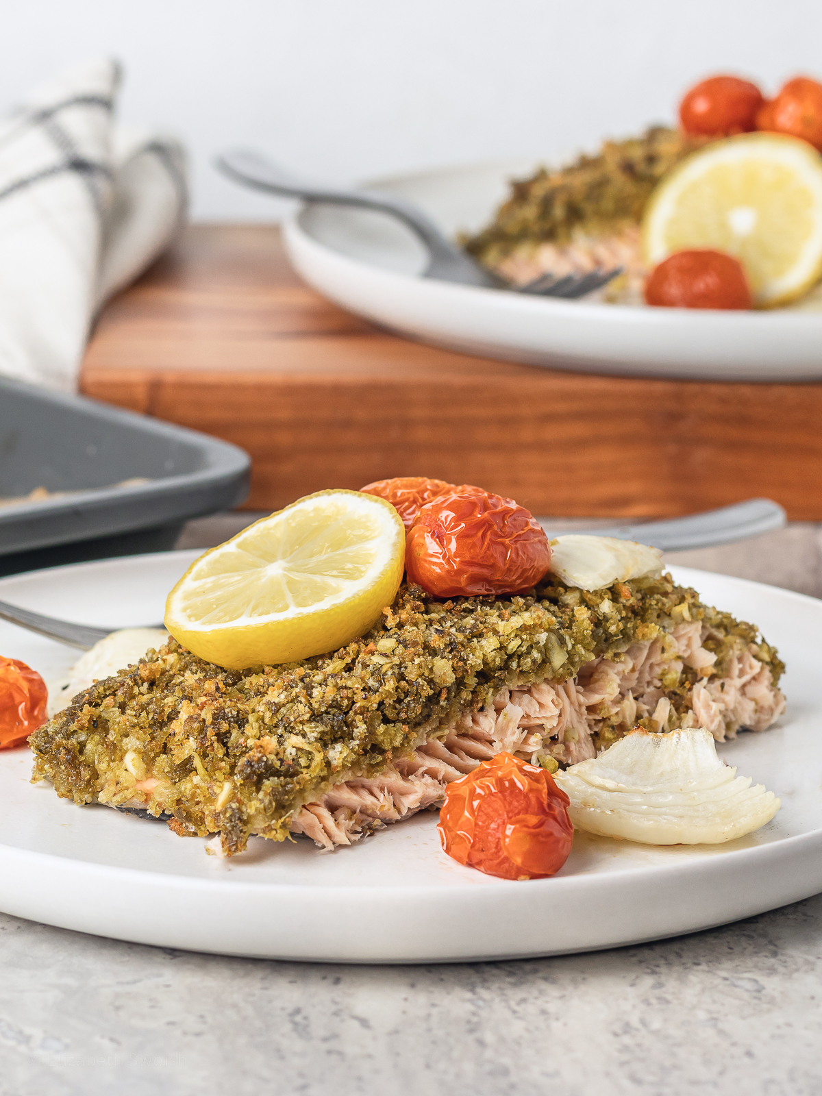 Two plates of Pesto Crusted Salmon with cherry tomatoes and onion. Garnished with lemon slices.