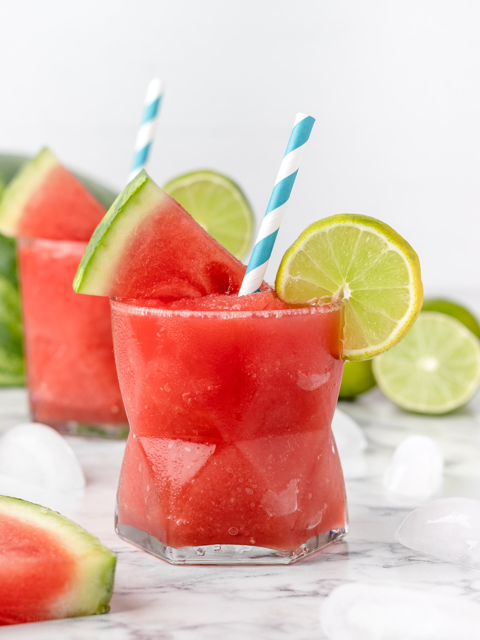 Two watermelon lime slush with blue striped straw, a lime slice, and a watermelon slice.
