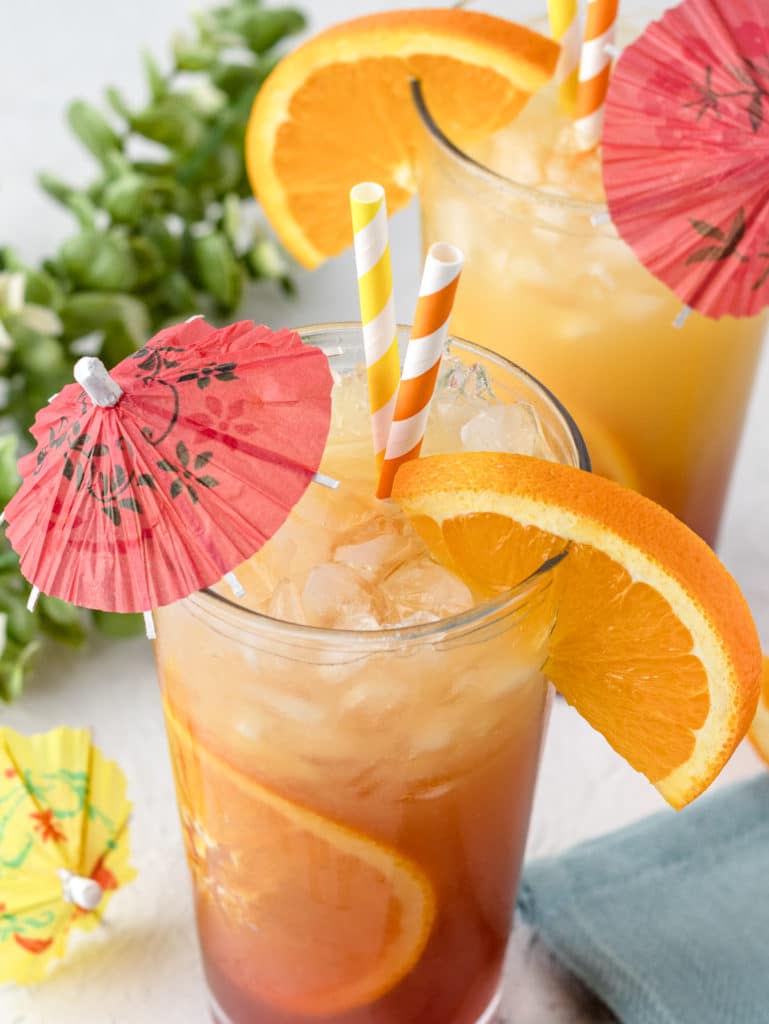 Sunrise Mocktail Spritzer in a tall glass garnished with a red umbrella, yellow and orange striped straws, and an orange slice.