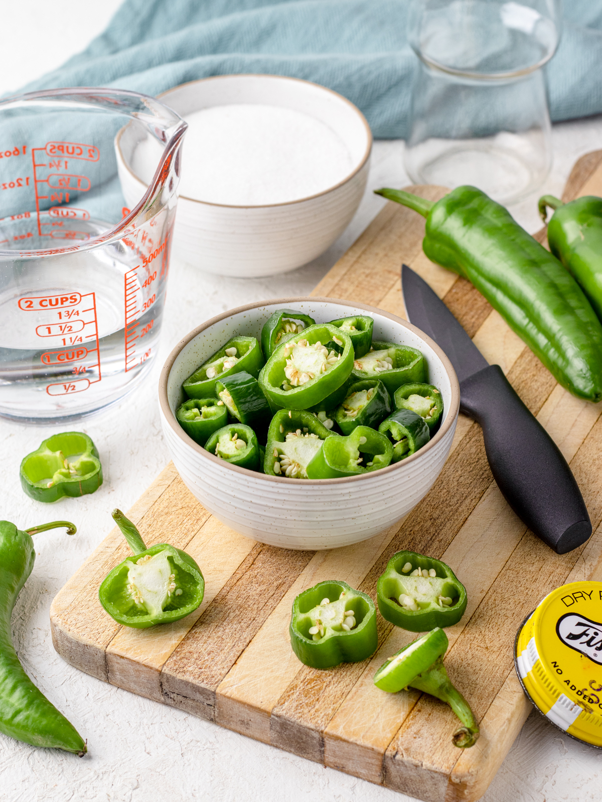 Bowl of sliced jalapenos setting on a cutting board with a knife, water, sugar, a jar for storing, and a blue towel on the side.