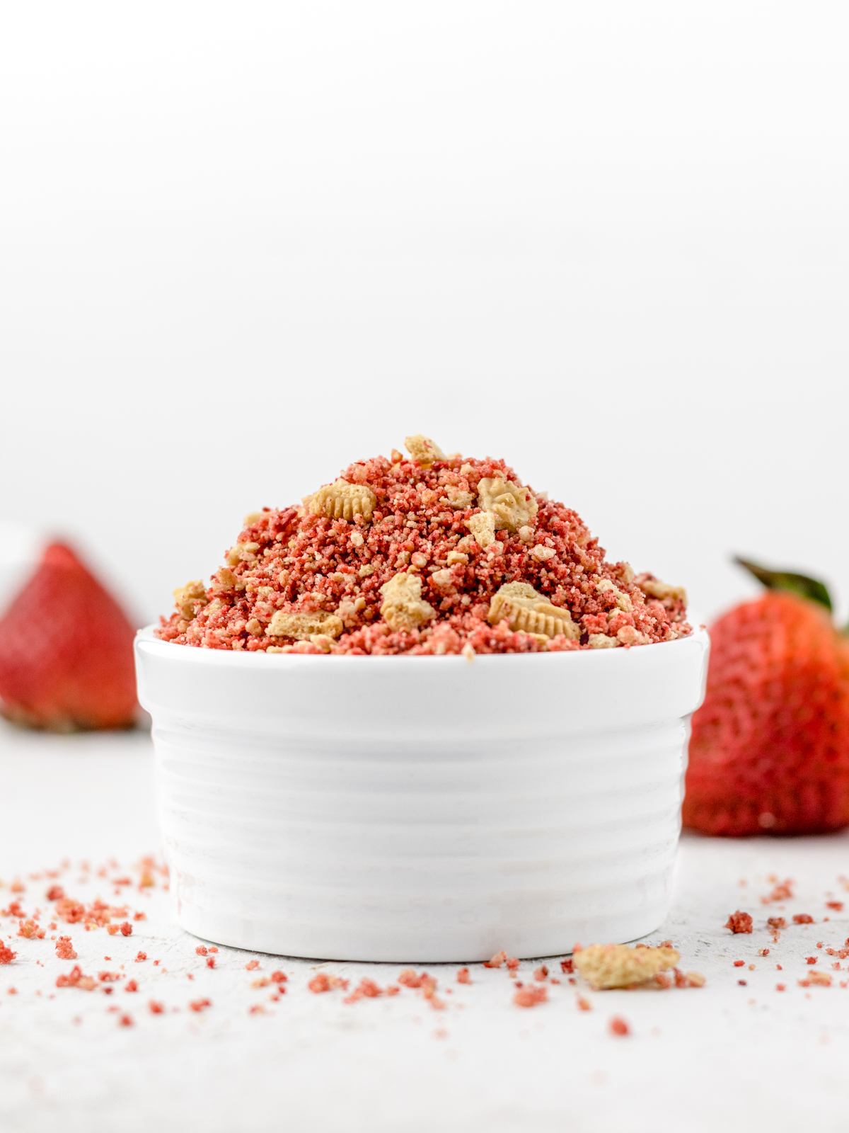 Strawberry Crunch Topping in a white bowl surrounded by cookie crumbs and strawberries.
