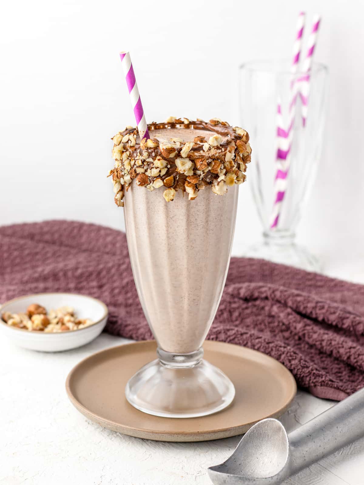 Nutella Milkshake in a milkshake glass with purple straw and glass rimmed with hazelnut spread and chopped hazelnuts.