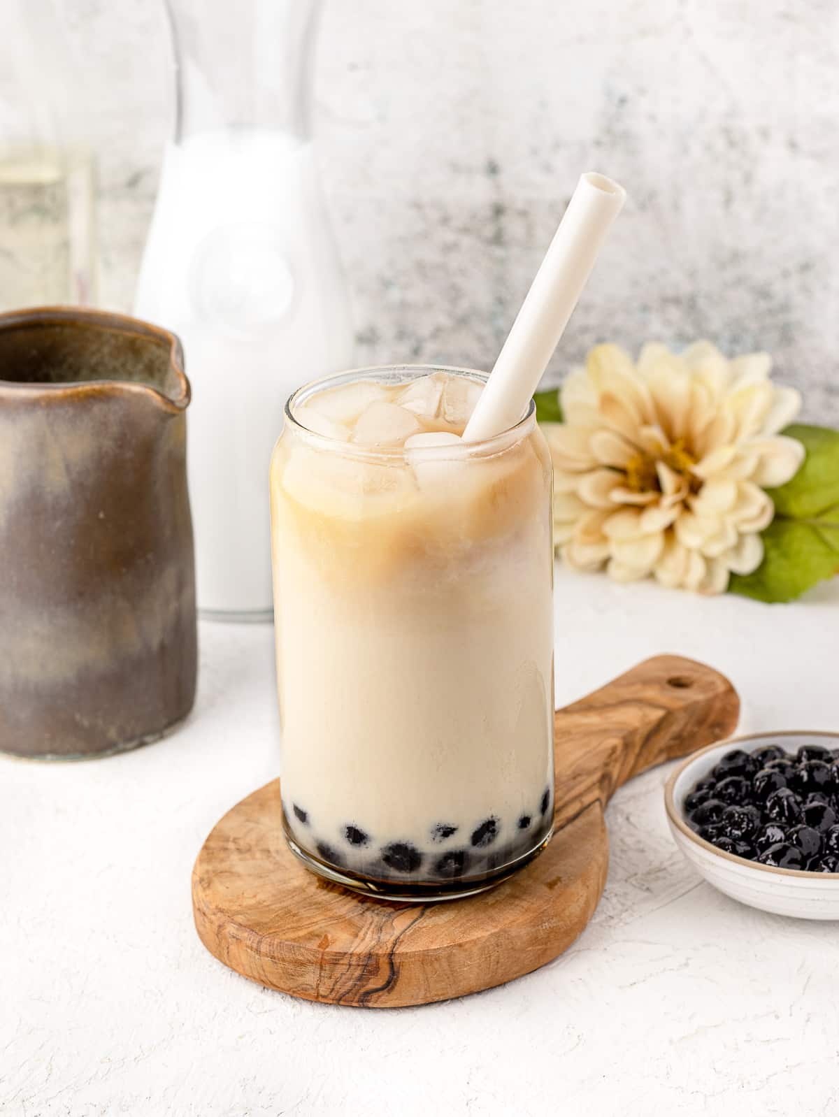 Earl Grey Milk Tea with boba. More bobas on the side, half and half in a jar, tea in a little pot, and a beige flower.