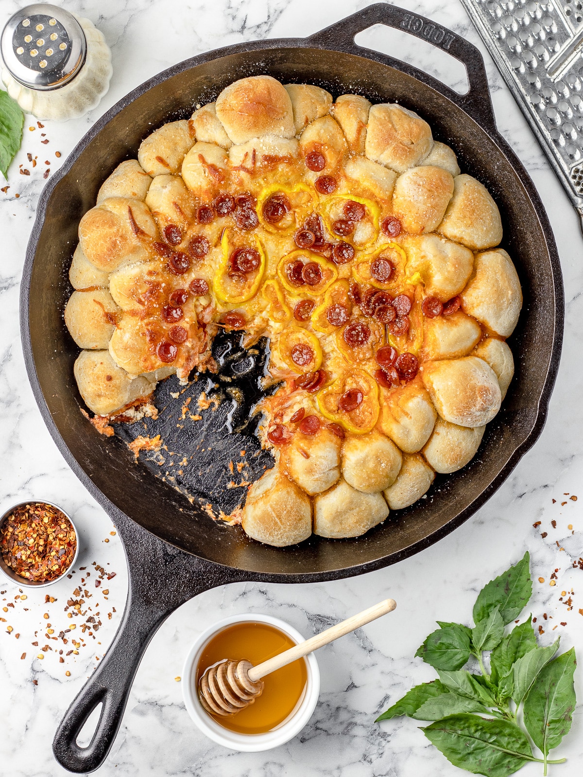 Pizza Dip with Pizza Dough Balls in a large cast iron skillet and partially eaten. It is surrounded by more honey, parmesan cheese, basil, and red pepper flakes.