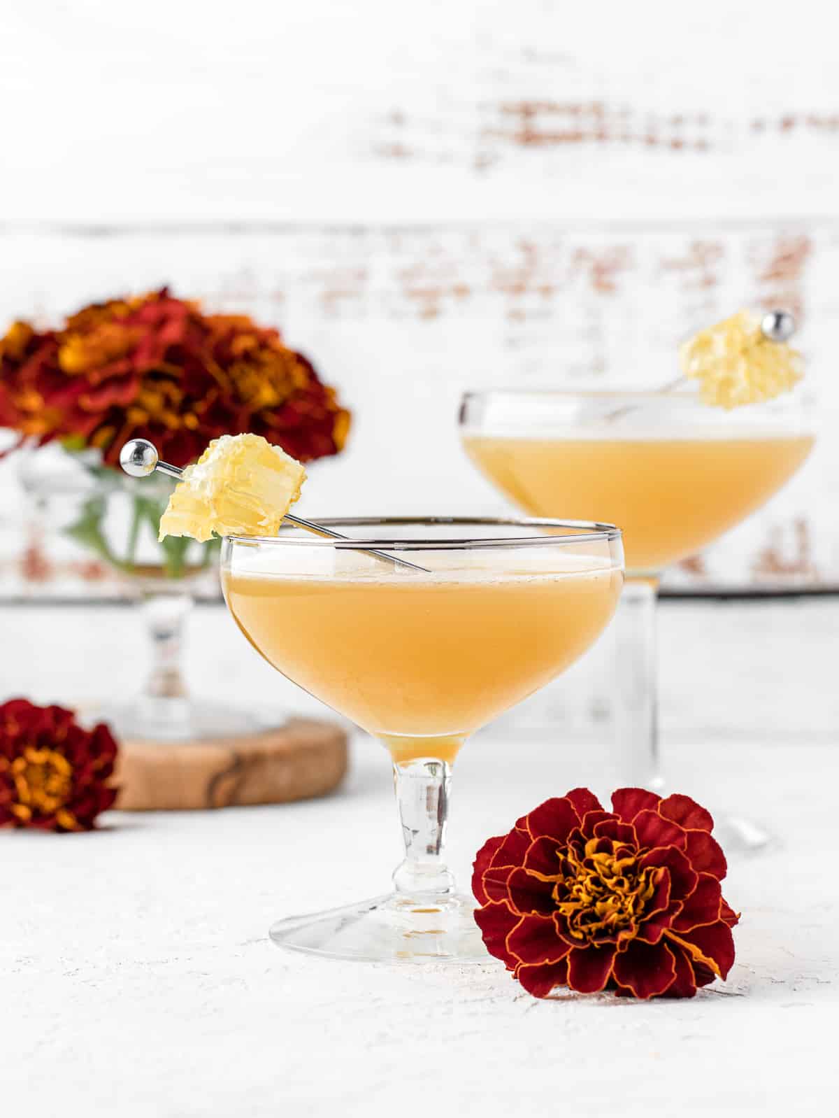 Two mocktails in coupe glasses with a cocktail pin of fresh honeycomb, surrounded by red and orange mums.