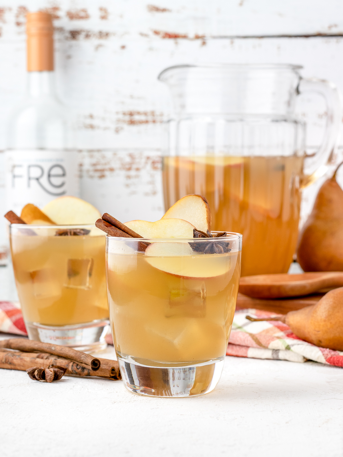 Scene of pitcher of Apple Cider Sangria Mocktails with two full glasses of it and a bottle of alcohol removed Moscato. Decorated with pears, apples, cinnamon sticks, and star anise.