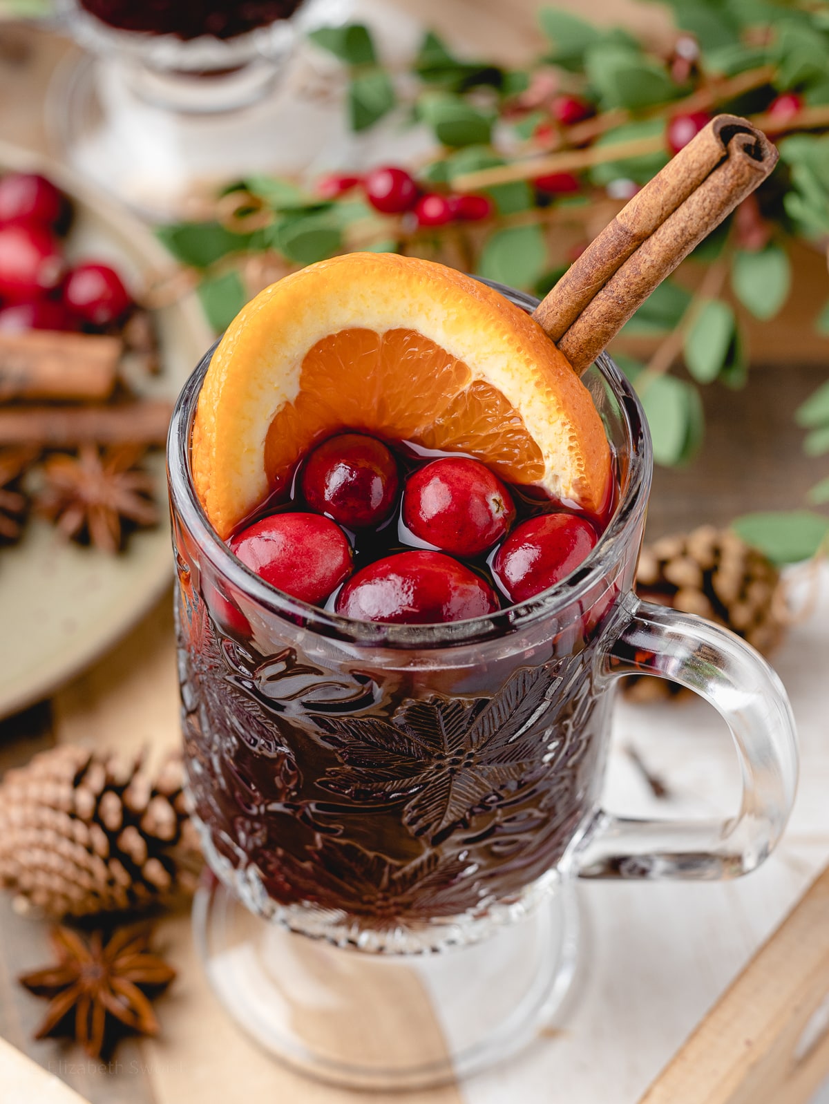 Non alcoholic mulled wine in a handled glass mug. Close up to see garnish of orange slice, cranberries, and cinnamon stick.