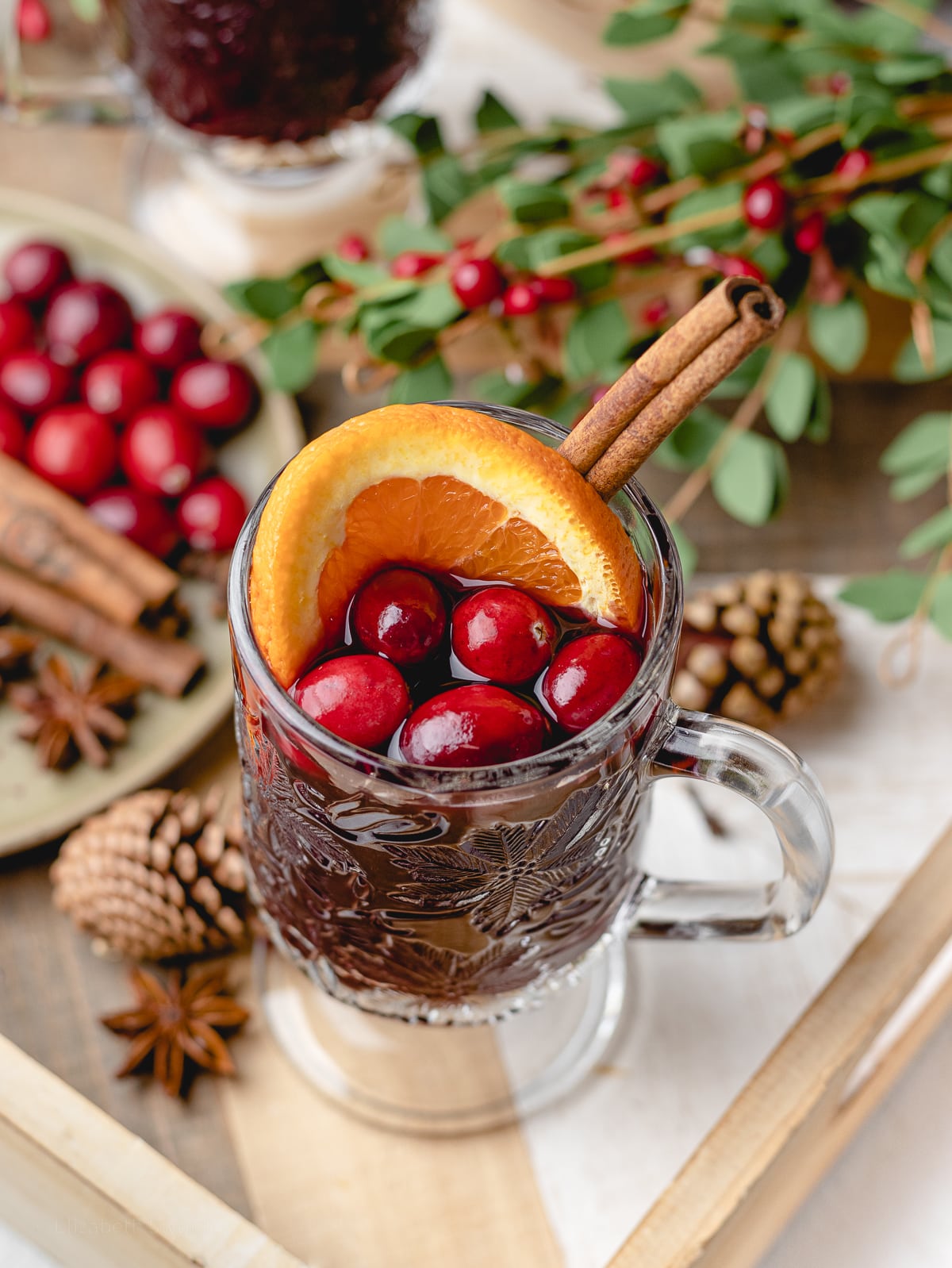 Alcohol free mulled wine in a handled glass mug. Close up to see garnish of orange slice, cranberries, and cinnamon stick.