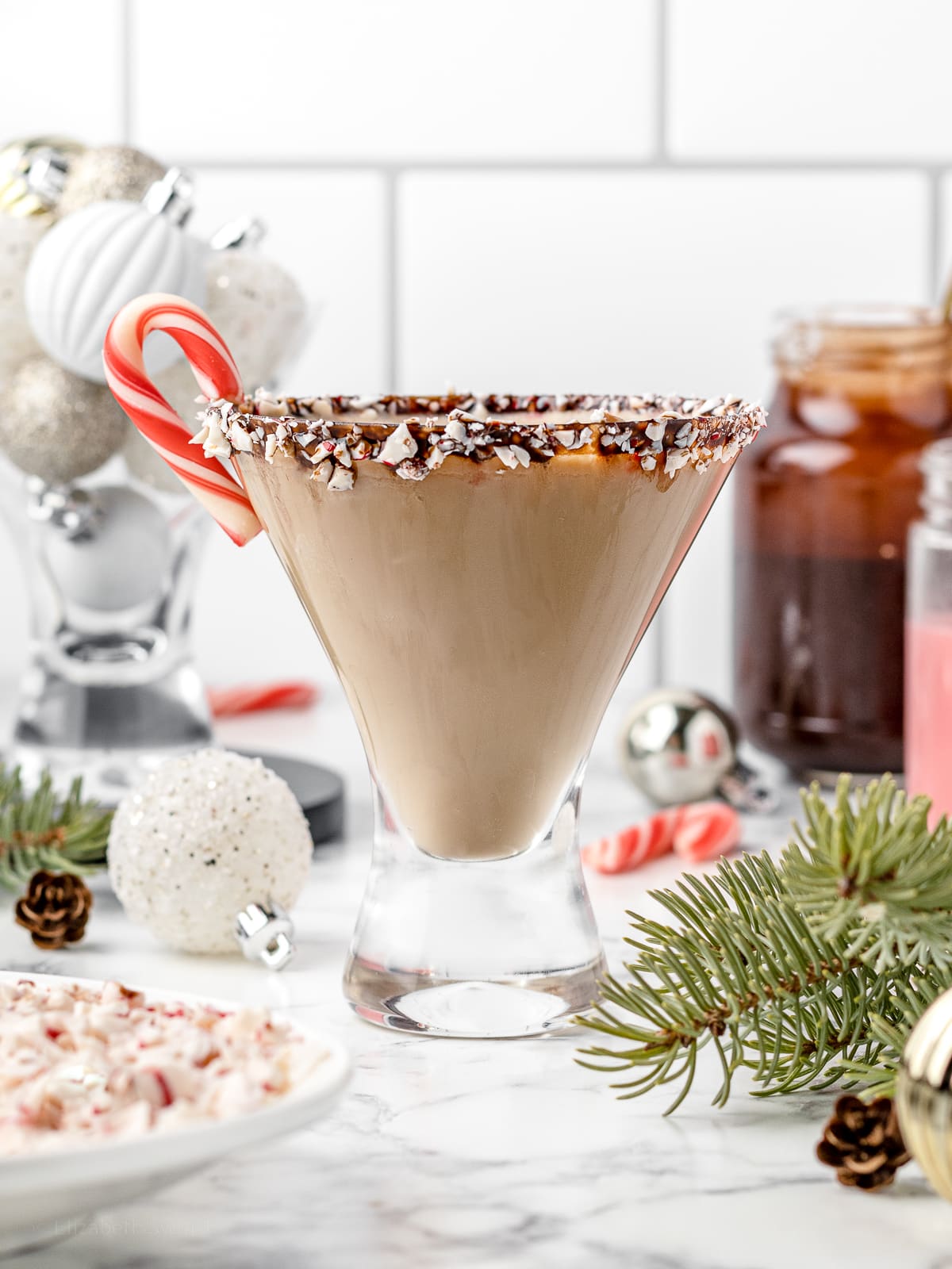 Peppermint White Russian Mocktail surrounded by crushed candy canes, chocolate syrup, peppermint syrup, candy canes, Christmas ornaments and pine trees