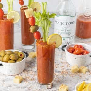 Basil Bloody Mary Mocktail and Cocktail recipe. Drinks surrounded by olives, lemon wedges, tomatoes, tajin.