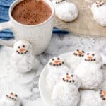 Snowman Cookies surrounded by mugs of hot cocoa and snowflakes.