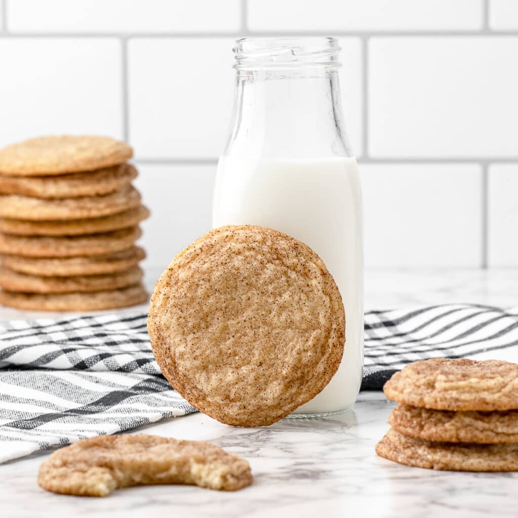 Stacks of snickerdoodles without cream of tartar, one snickerdoodle with a bite taken from it, and another snickerdoodle leaned up against a jar of milk.