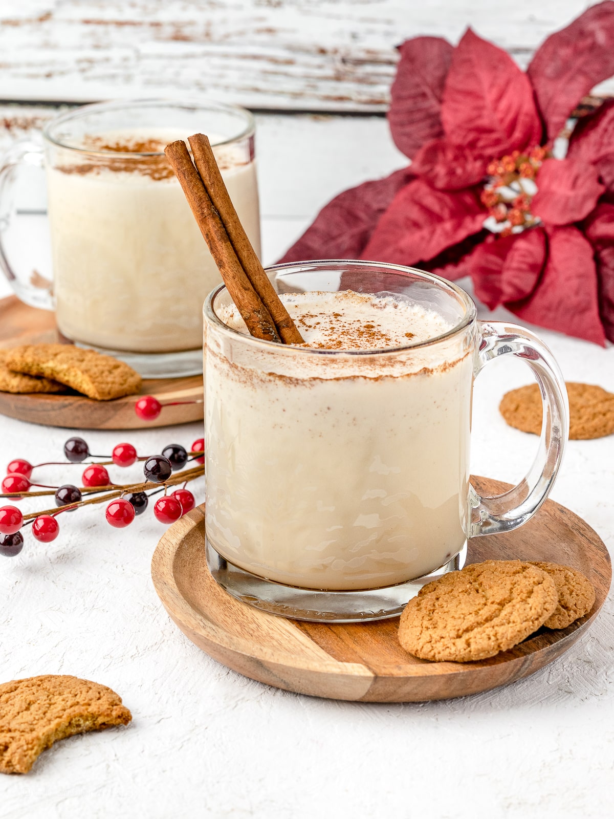 Two mugs of cold coquito garnished with ground cinnamon and a cinnamon stick. Ginger cookies served on the side.