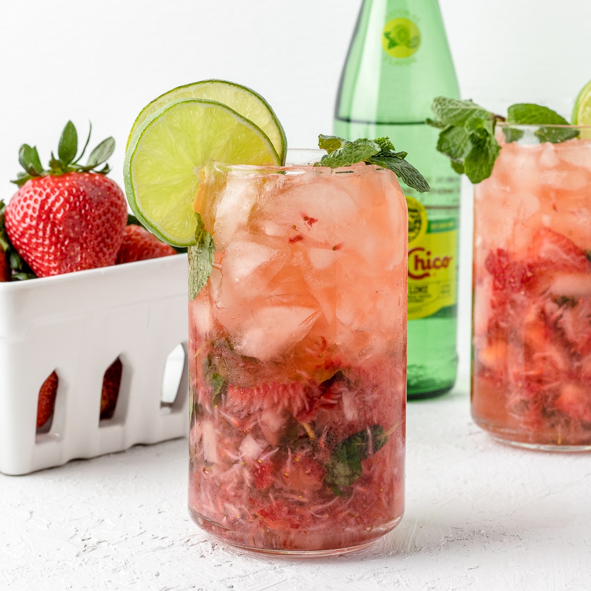 2 non alcoholic strawberry mojito mocktails garnished with lime slices and fresh mint leaves. Fresh strawberries and mineral water in the background.