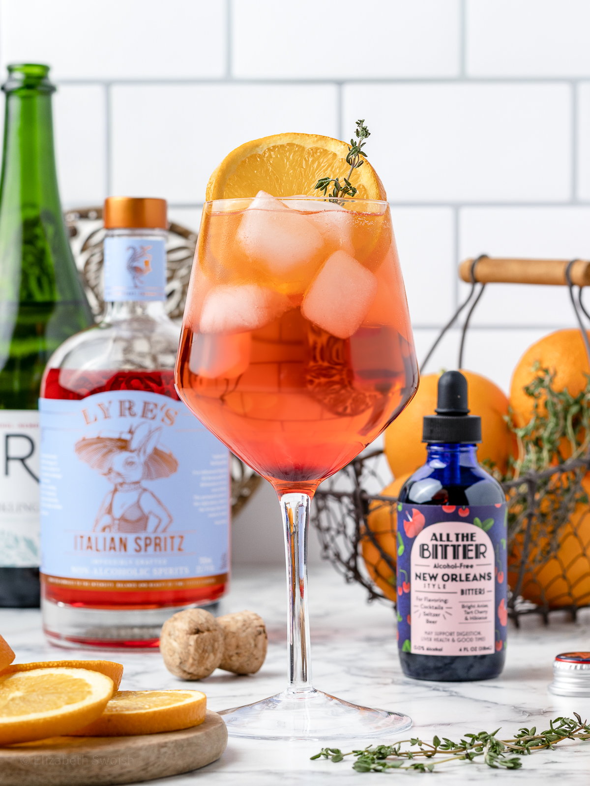 An orange and red Aperol Spritz Mocktail surrounded by the ingredients used to make one.