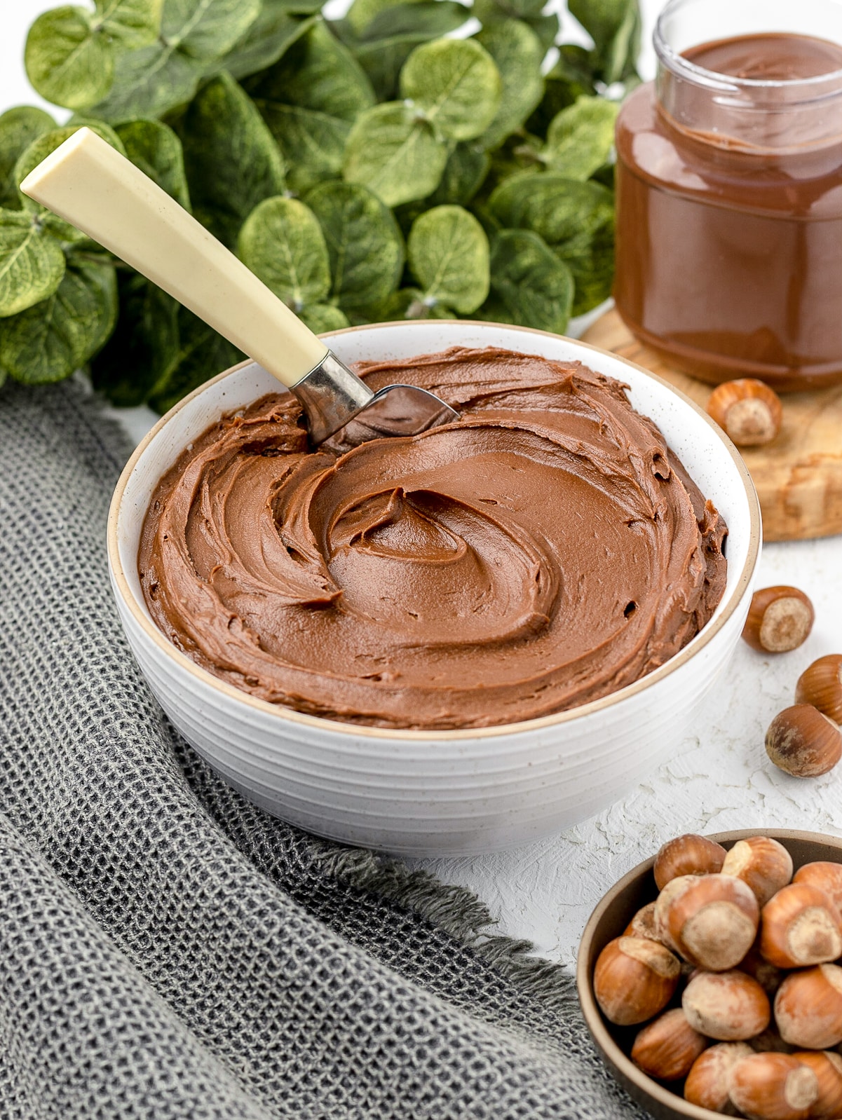 Nutella frosting in a bowl with a metal spreading tool. Whole hazelnuts and Nutella in the background.