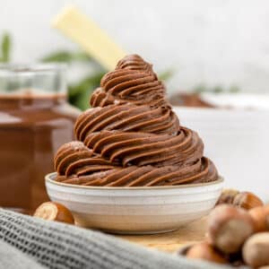 Swirls of Nutella Buttercream Frosting in a small dish. Surrounded by whole hazelnuts and Nutella.
