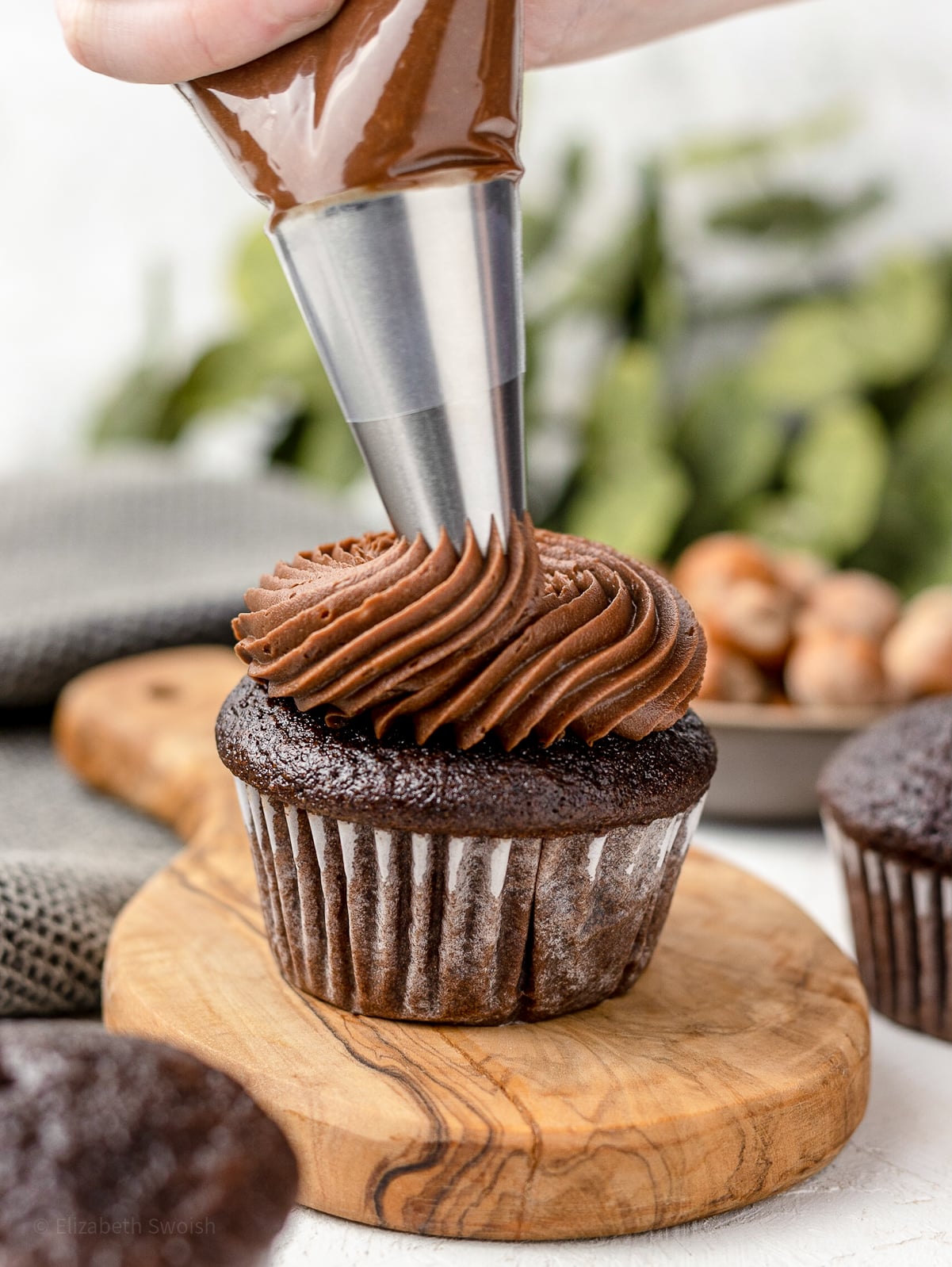 A hand holding a piping bag while piping Nutella Buttercream Frosting on a chocolate cupcake.