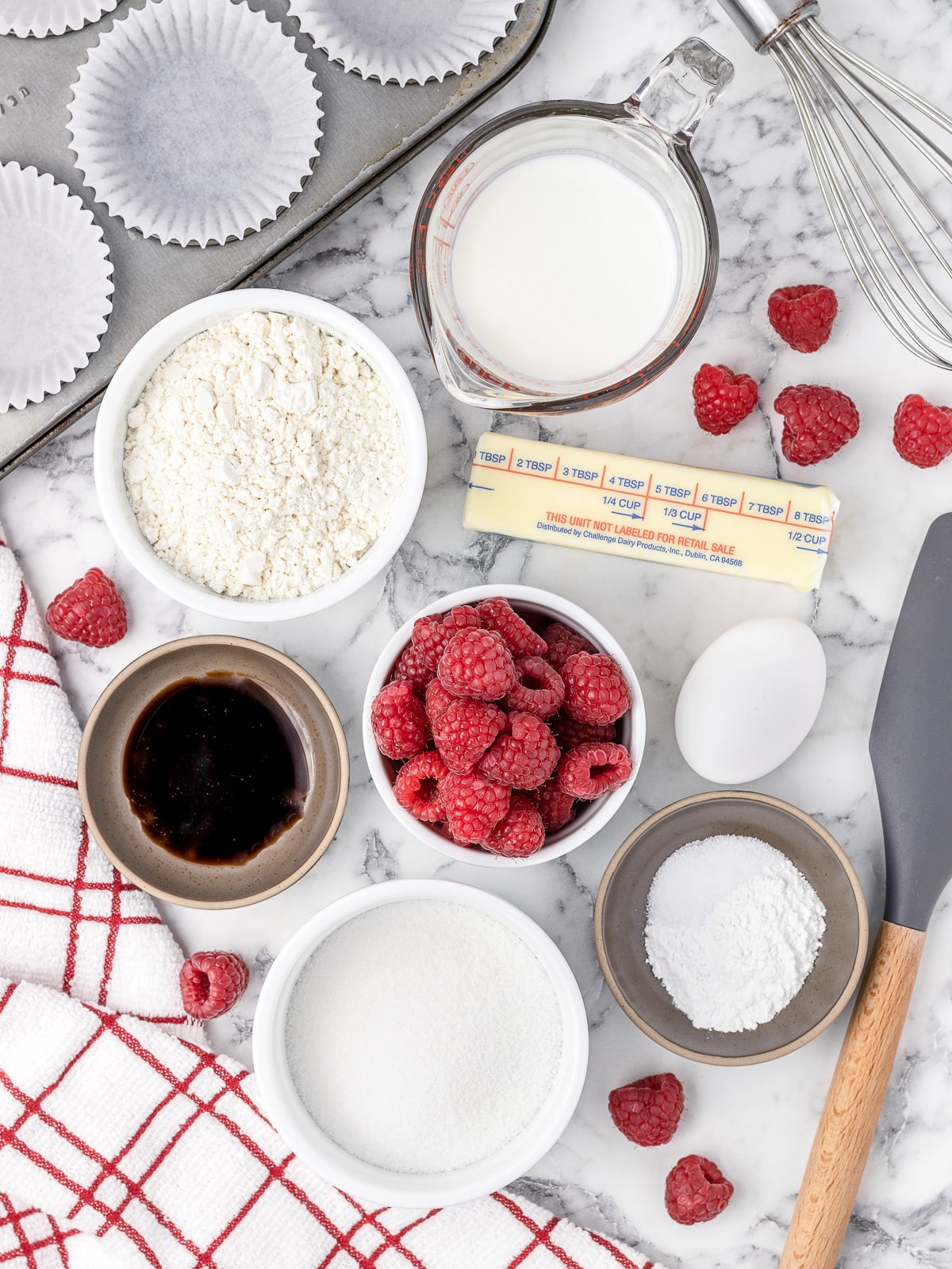 All the ingredients to make Raspberry Cupcakes with cupcaking baking pan lined with baking cups, whisk, spatula, and kitchen towl.