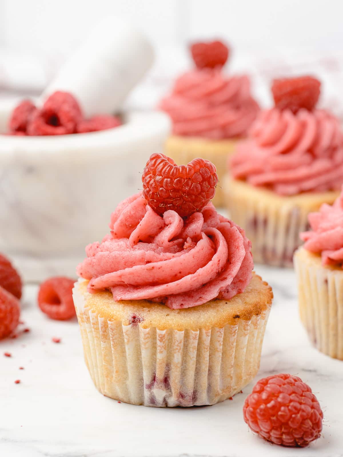 Decorated cupcakes with freeze dried raspberries in a mortar and pestle with fresh raspberries scattered.