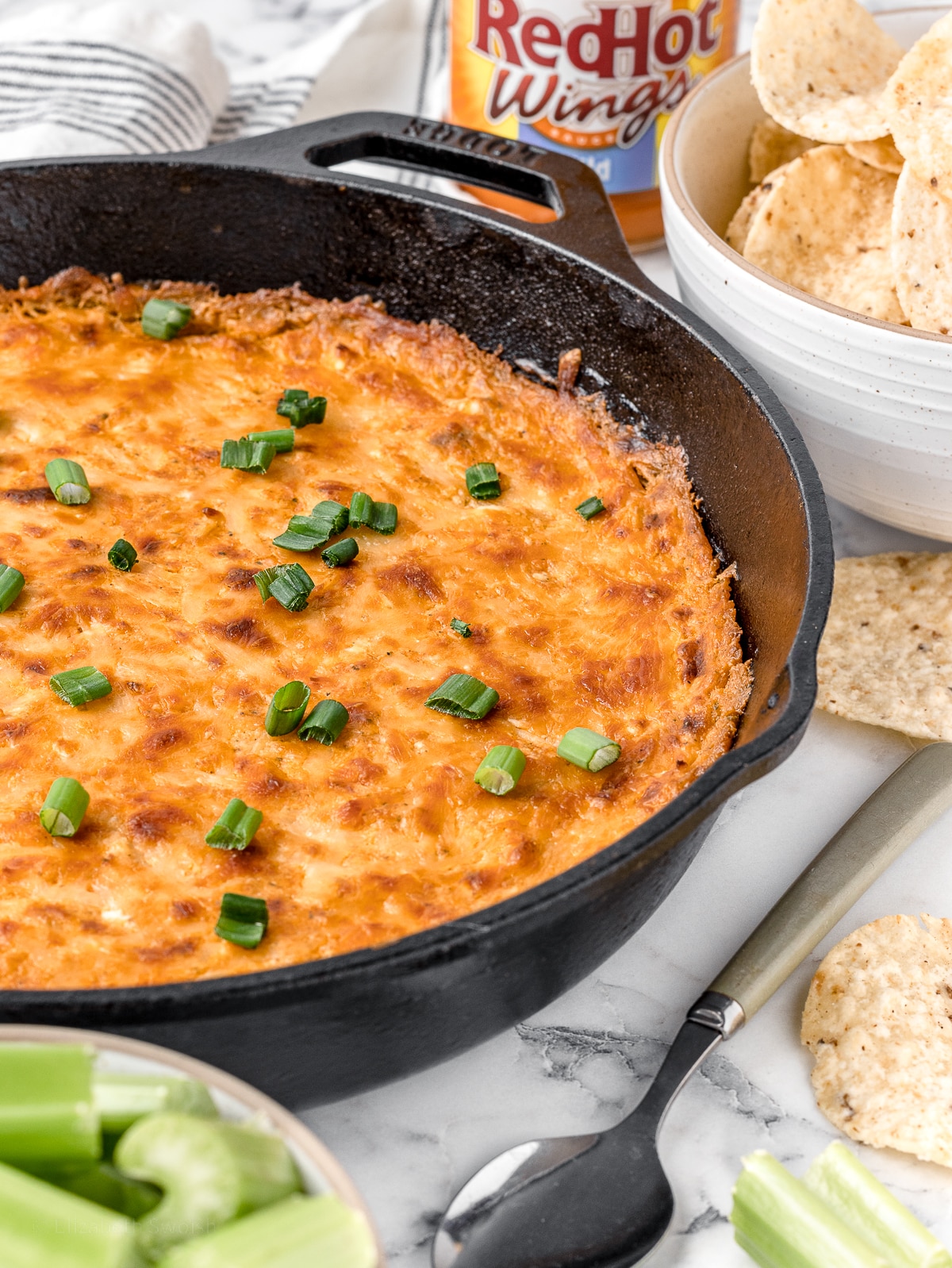 Smoked Buffalo Chicken Dip garnished with freshly chopped green onions. Celery, tortilla chips, and a spoon on the side for serving.