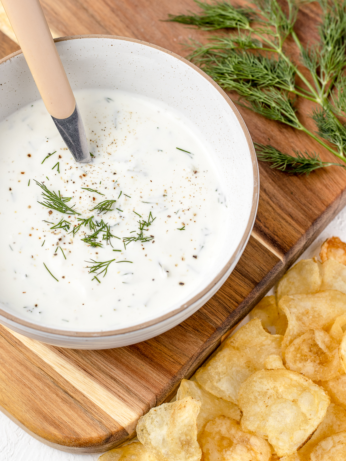 Bowl of Yogurt Dill Sauce with a spoon, dill and potato chips on the side.