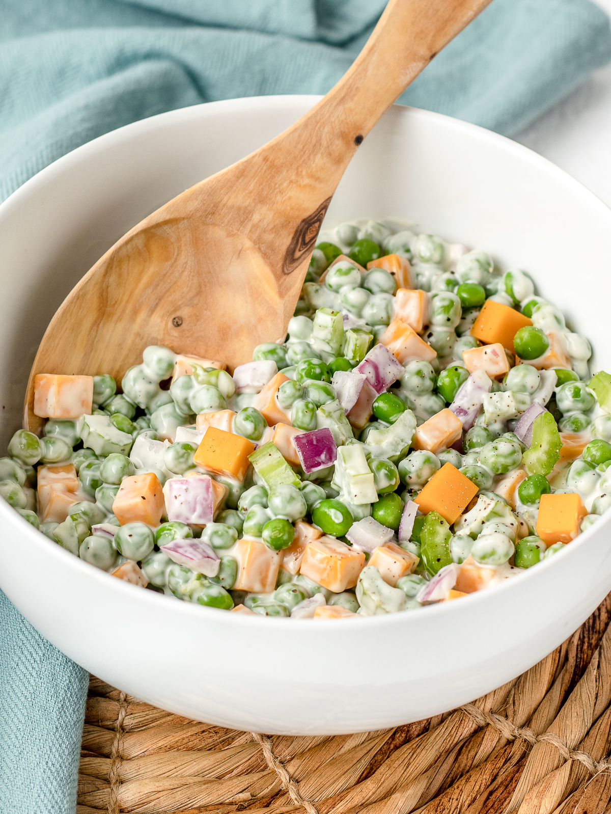 Spoon in a large bowl of colorful pea salad- green peas and celery, purple red onion, and orange cheddar cheese.