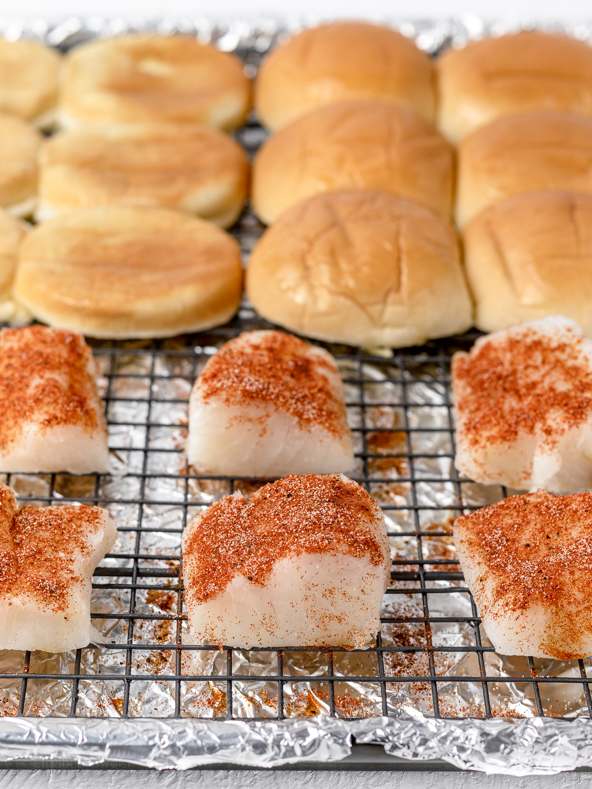 Seasoned cod fish and slider buns on a rack lined baking sheet ready to go in the oven.