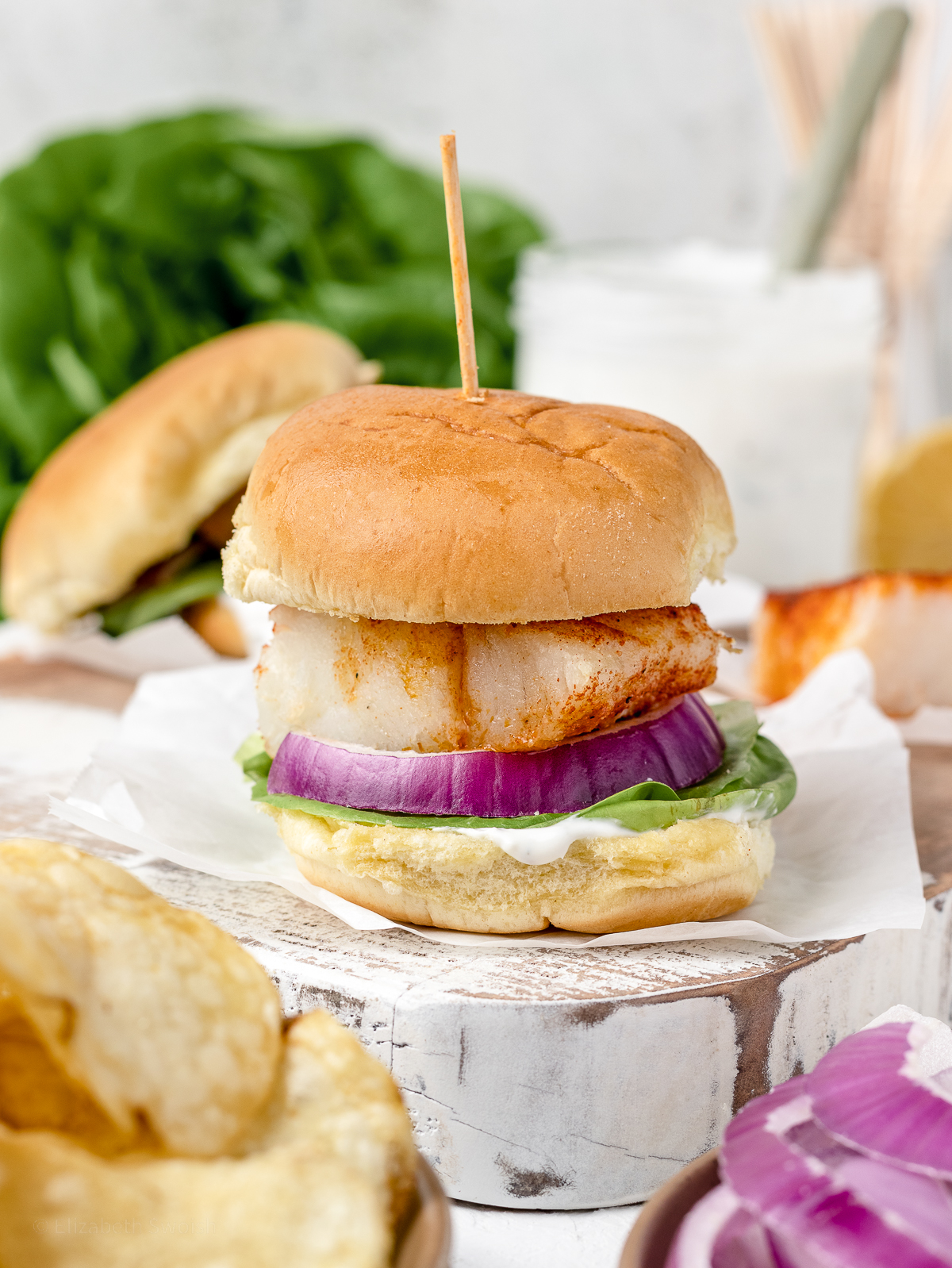 Baked Cod Fish Sliders piled high with lettuce, red onion, and yogurt dill sauce. Potato chips and more sauce served on the side.