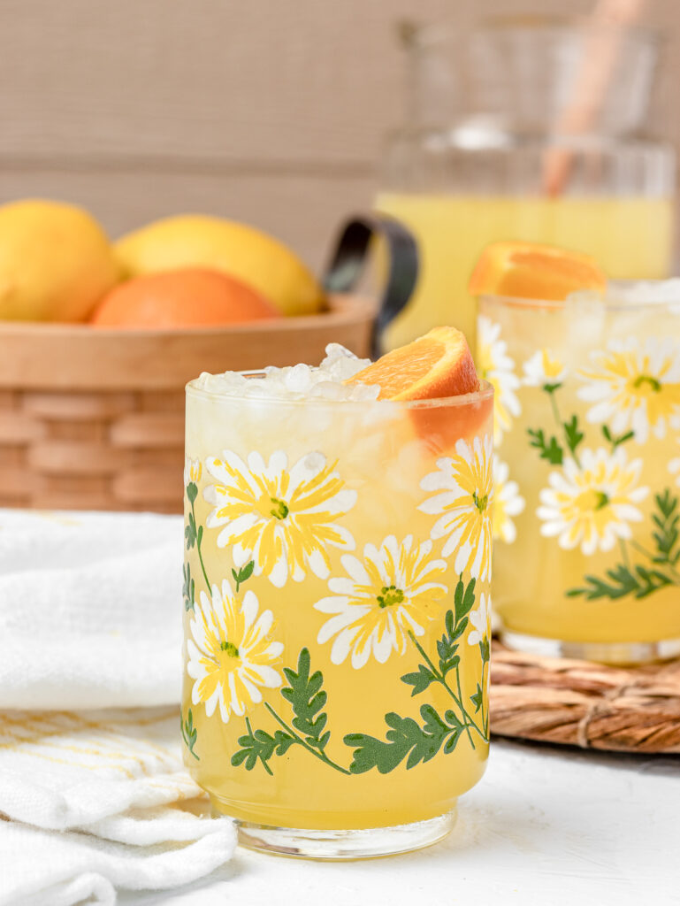 Two glasses of Orange Lemonade with a large pitcher of more in the background and lemons and oranges on the side.