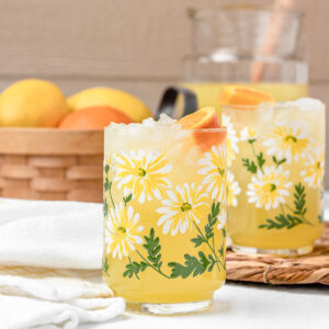 Two glasses of lemonade with a large pitcher of more in the background and lemons and oranges on the side.