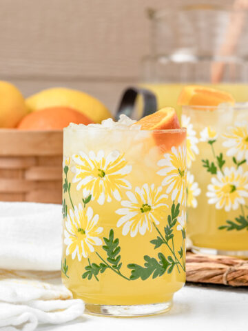 Two glasses of lemonade with a large pitcher of more in the background and lemons and oranges on the side.
