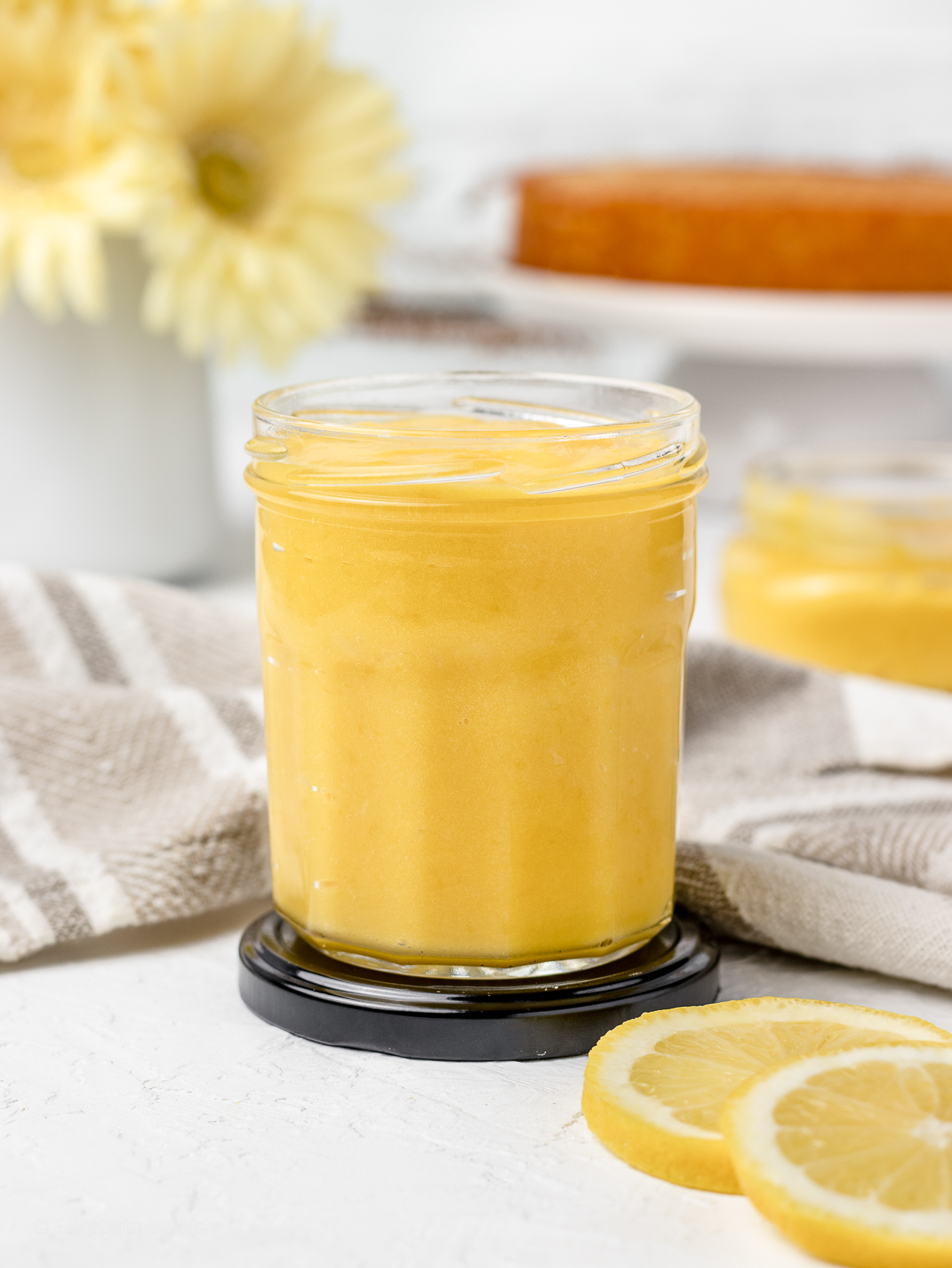 Cooked and cooled lemon curd in a jar with baked lemon cake in the background.