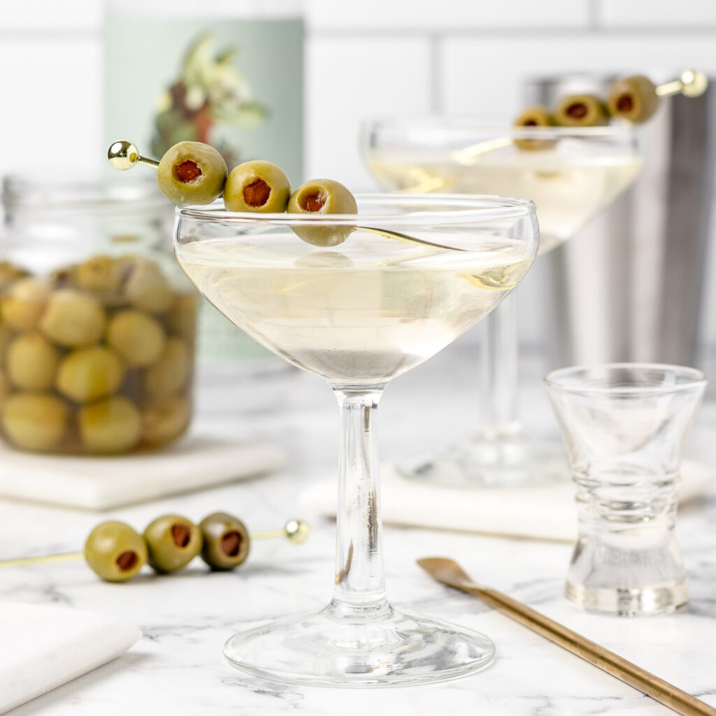 2 Non Alcoholic Dirty Martini Mocktails with a stirrer, shot glasses, non alcoholic spirits, and olives in the back ground.
