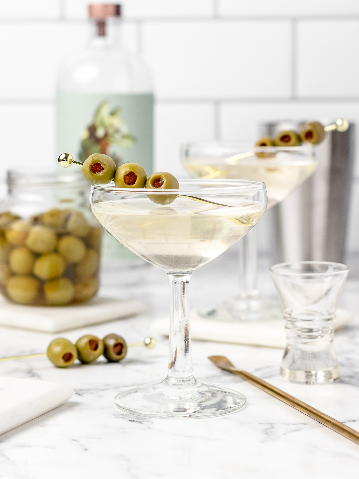 Non Alcoholic Dirty Martini Mocktail with stirrer, shot glasses, non alcoholic spirits, and olives in the back ground.