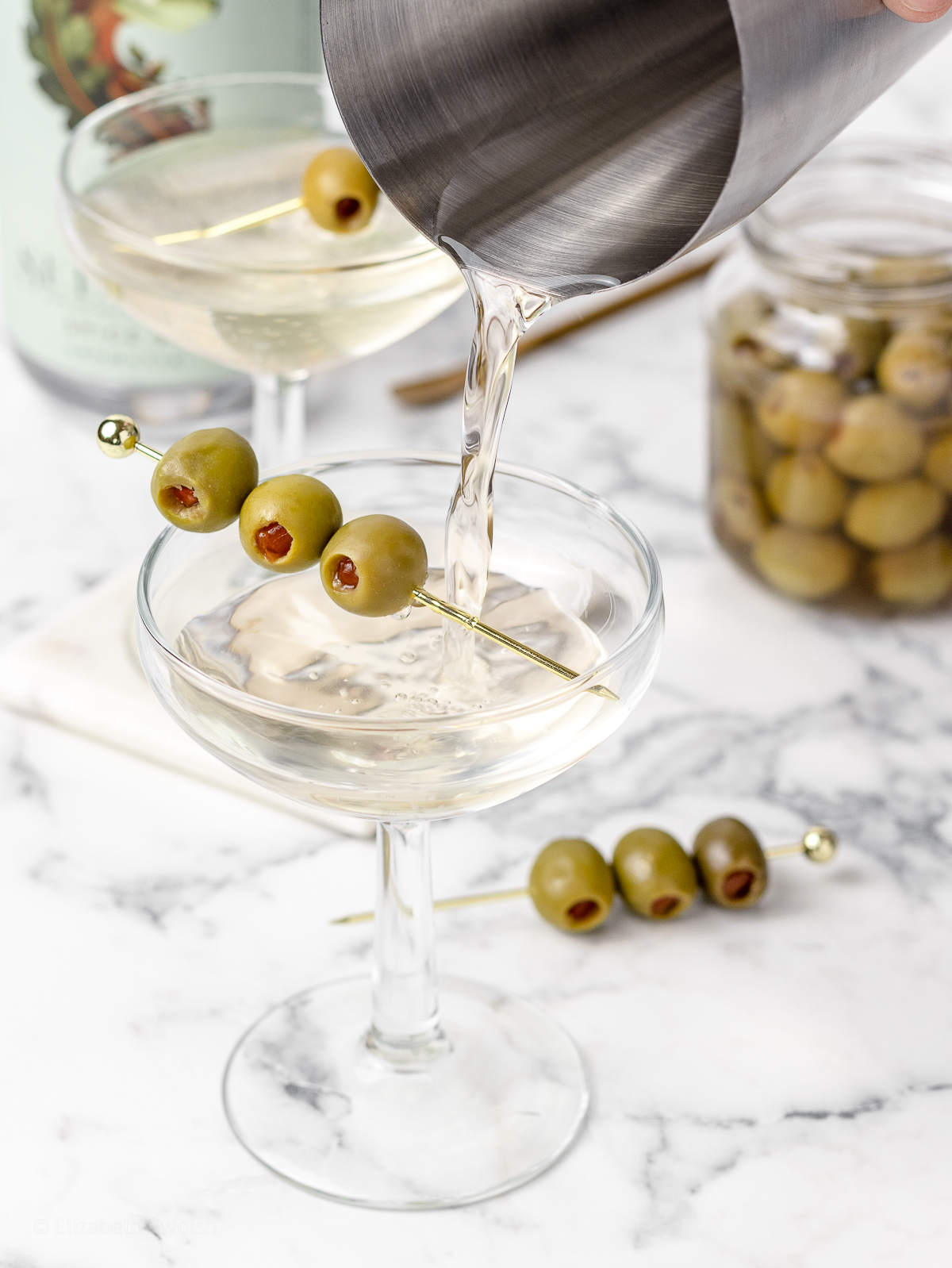 Pouring into a glass garnished with 3 olives and more olives on the side.