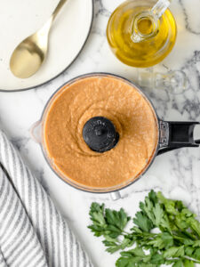Fully blended hummus in a food processor with a serving plate, parsley, and extra olive oil on the side.